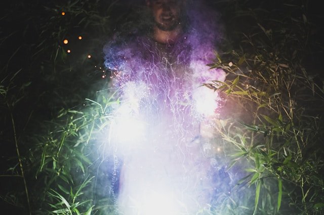 A magician in a dark, forested space with twinkling lights in the background, holding bright sparklers producing purple and violet smoke.