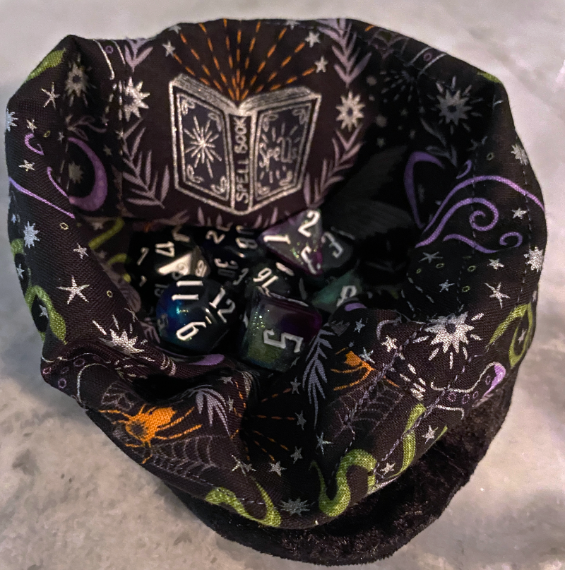 IMG_5916_refined Photo of a dice bag resting on a quartz table with a set of dice nestled inside. The bag is black velvety fabric on the outside and spellbound themed print on the inside. The print of a spell book outlined in silver is visible, as is bits of green, orange, and purple objects like snakes and spiders.