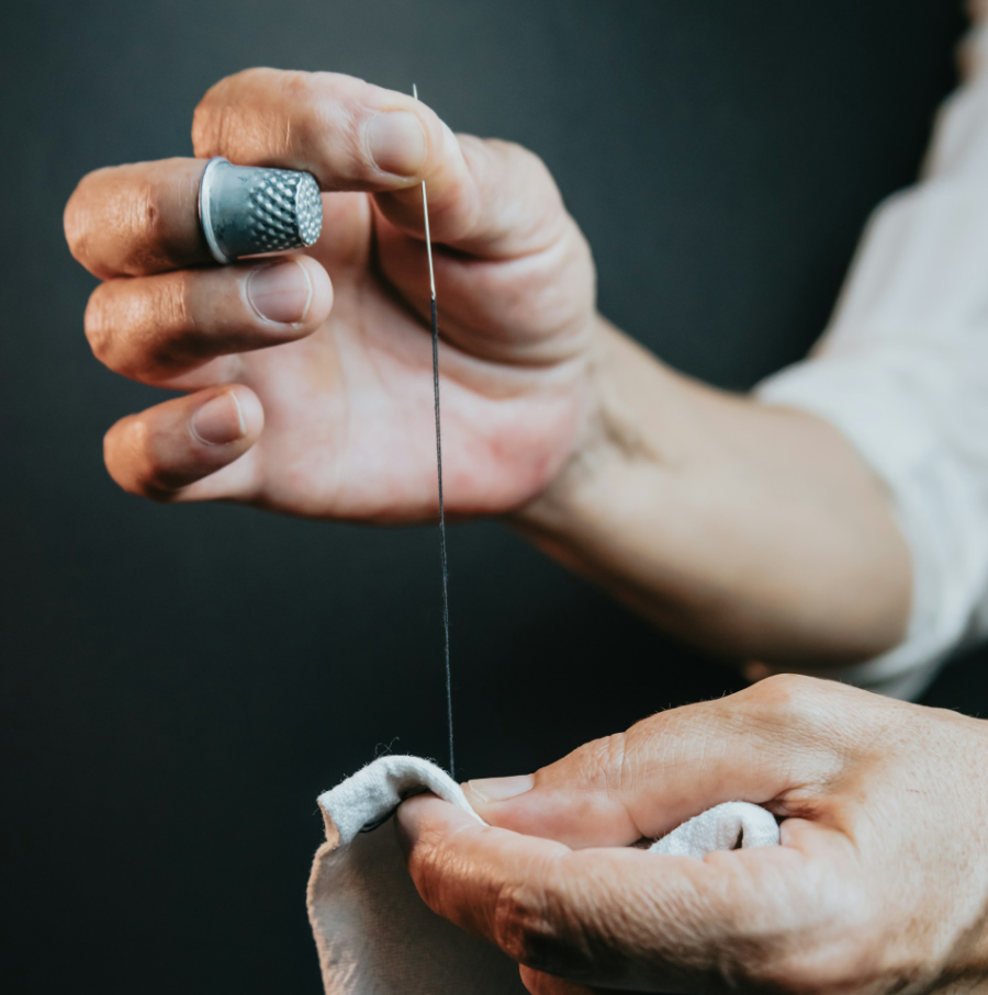 Close up photo of hand sewing. The left hand is holding a white fabric while the right holds a needle upright, with a dark thread leading to the fabric. The right hand has a thimble on one finger.